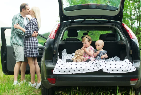 Children sitting in car trunk. Parents stand nearby and kiss.