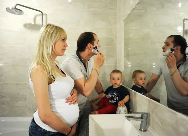 The father costs in a bathroom and shaves the person. Pregnant mother and the kid watch the father.