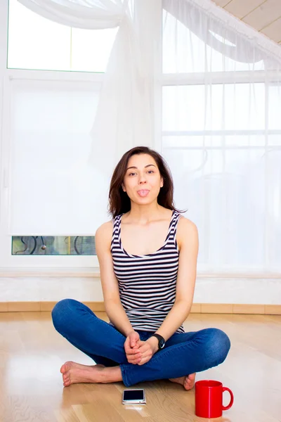 Pretty young woman dressed in jeans and a T-shirt  sitting on the floor at home sticking out his active tongue
