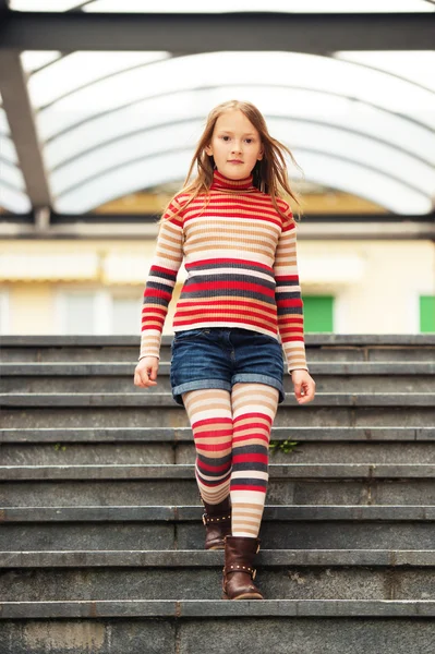 Fashion portrait of a cute little girl in a city, wearing brown boots, denim shorts, stripes rollnecck pullover and tights