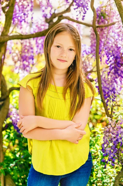 Outdoor stylish vertical portrait of a cute little girl of 8-9 years old, standing next to beautiful purple wisteria flowers, wearing green blouse, arms crossed