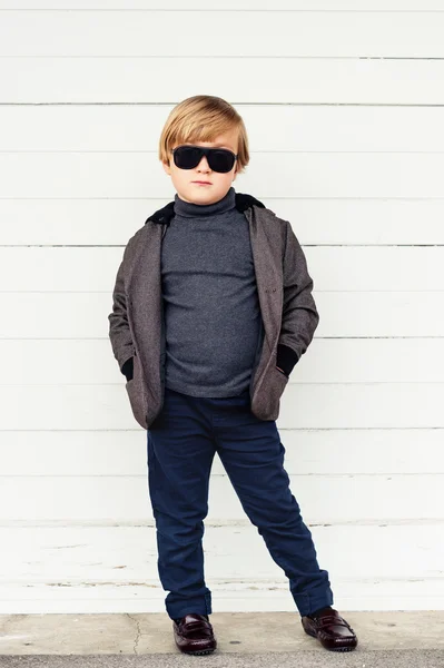 Fashion kid in sunglasses, cute little boy wearing dark jacket, trousers, roll neck t-shirt and moccasins, standing against white wooden background