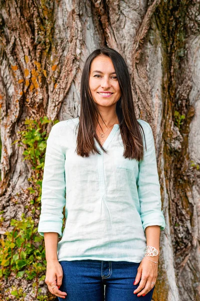 Stylish cheerful brunette leaning on a tree in a park on a sunny day, wearing denim jeans, mint blouse