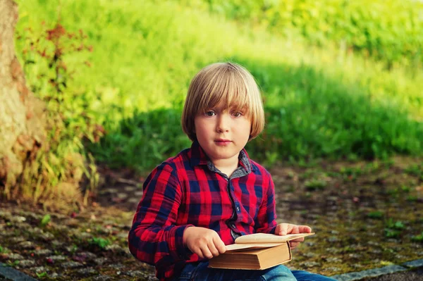 Sweet little boy reading old book in park, wearing red and blue plaid shirt. Toned image