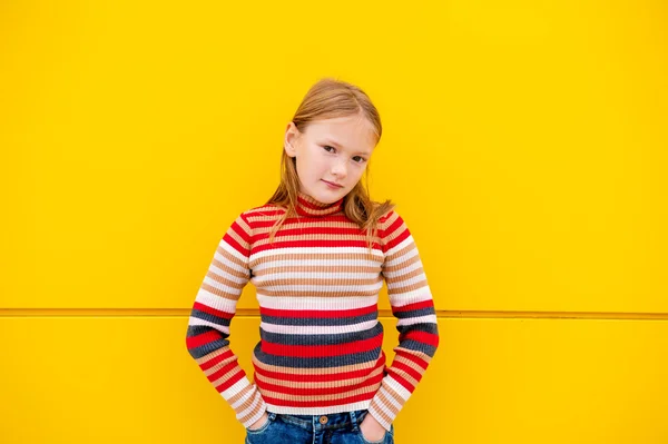 Portrait of a cute little girl of 8 years old, wearing stripe roll neck pullover and denim skirt, standing next to bright yellow wall