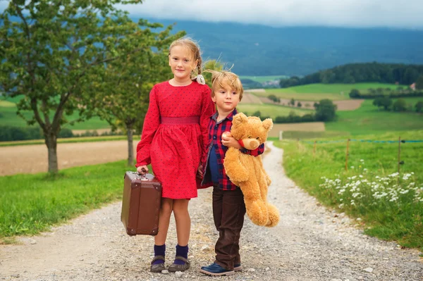 Two cute kids, little girl and her brother, walking down the small road, holding big teddy bear and old brown suitcase