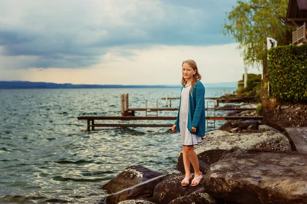Adorable little girl of 8-9 years old playing by the lake, wearing sandals, dress and blue knitted jacket
