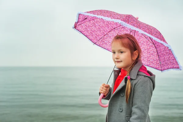 Pretty little girl under the rain, wearing grey coat, holding pink umbrella, standing by the lake