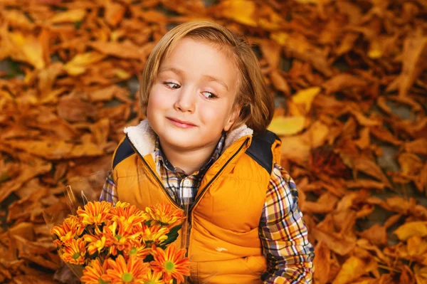 Autumn portrait of adorable little blond boy of 4 years old, wearing warm yellow vest coat, holding small bouquet of orange chrysanthemum flowers