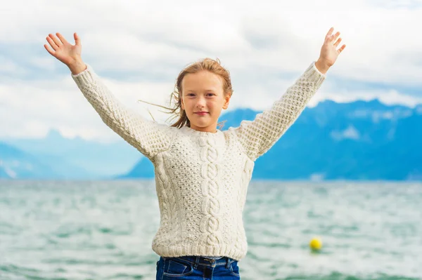 Cute little girl of 8 years old playing by the lake on a very windy day, wearing warm white knitted pullover, arms wide open