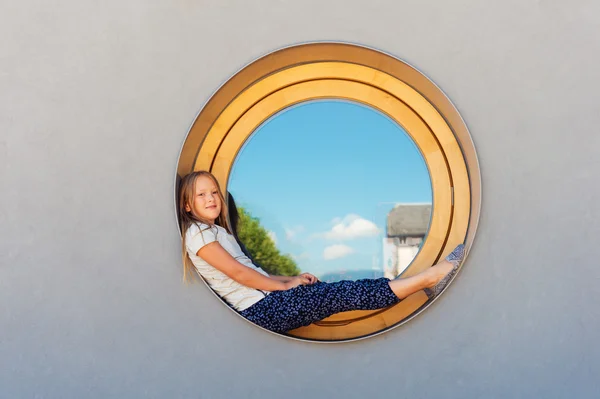 Funny little girl sitting on a round window outdoors