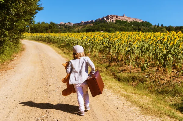 Outdoor portrait of a funny little boy walking down the road in countryside, holding old small suitcase and teddy bear toy, back view. Image taken in Tuscany, Italy