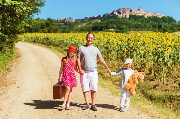 Outdoor portrait of a funny little kids and the father walking down the road in countryside, holding old small suitcase. Image taken in Tuscany, Italy