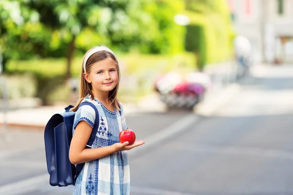 Pretty little 9 year old girl walking back to school, wearing blue vintage backpack, holding red apple
