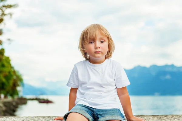 Portrait of beautiful smiling cute little boy. 3-4 years old little child playing outside by the lake Geneva, in summer or spring. Boy sitting alone by the lakeside at sunset, wearing white t-shirt