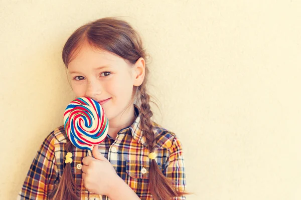 Portrait of a cute little girl of 7 years old, holding big colorful candy, wearing plaid shirt, toned image