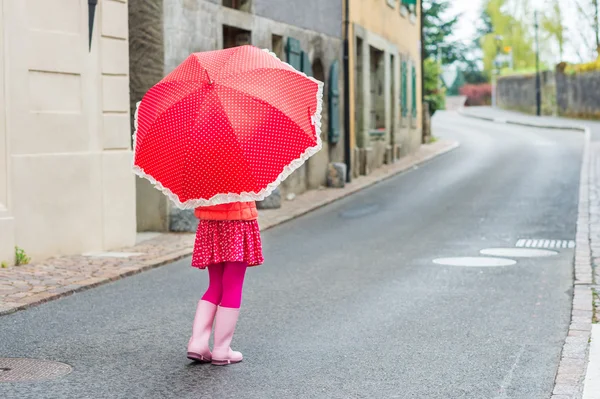 Cute little girl walking down the street, wearing pink rain boots, holding polka dot red umbrella, back view