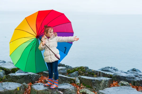 Outdoor portrait of a cute little girl of 6 years old with big colorful umbrella, playing by the lake on a cold rainy day, wearing warm white coat and rainbow boots