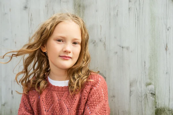 Close up portrait of a cute little girl of 8 years old with curly hair, wearing terracotta pullover, sitting against grey wooden background