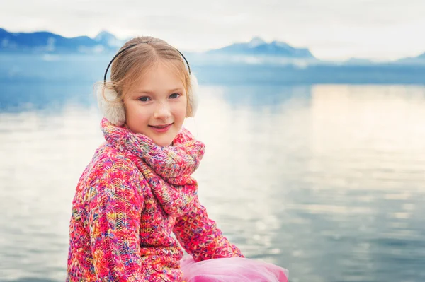 Outdoor close up portrait of adorable little kid girl of 8 years old resting by the lake Geneva on a nice winter day, wearing colorful pink pullover and warm earmuffs