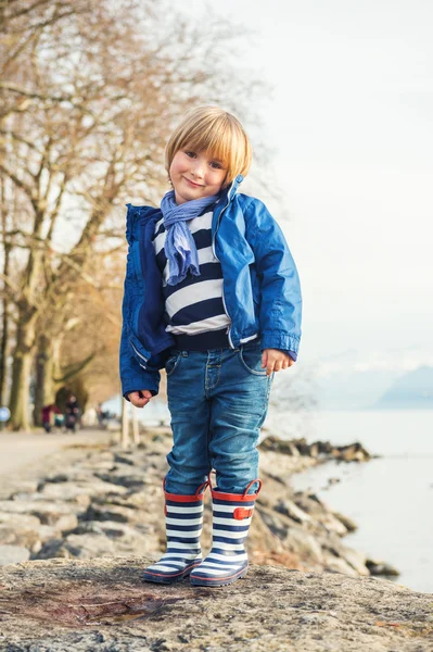 Outdoor portrait of adorable little blond boy of 4-5 years old, having fun by the lake on a nice sunny spring day, wearing warm blue jacket, scarf, denim jeans and stripes rain boots