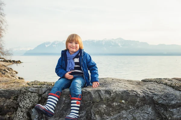 Outdoor portrait of adorable little blond boy of 4-5 years old, having fun by the lake on a nice sunny spring day, wearing warm blue jacket, scarf, denim jeans and stripes rain boots