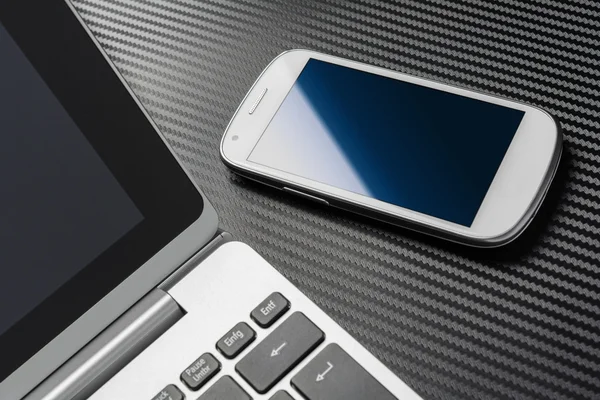 Business Work With White Blank Smartphone With Blue Reflection Lying Right To A Notebook Keyboard, All Above A Carbon Layer