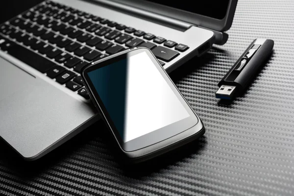 Blank Black Business Smartphone With Blue Reflection Leaning On A Notebook Keyboard Next To An USB Storage Flash Drive, All Above A Carbon Layer
