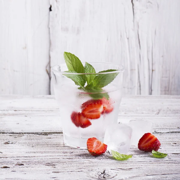 Glass of refreshing ice water with slices  juicy ripe strawberries, mint leaves and  cubes in a simple white wooden background. The concept  summer vacation relaxation