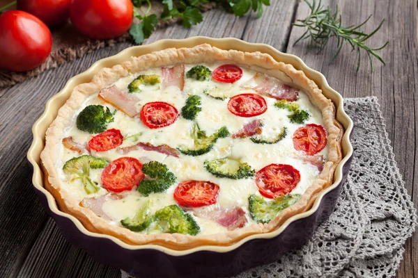 Traditional french homemade quiche lorraine tart pie recipe with broccoli, bacon, cheese and tomatoes in baking dish on vintage cloth.