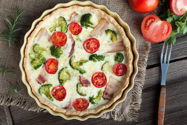 Quiche lorraine traditional french homemade tart pie with bacon tomatoes cheese and broccoli in baking dish on vintage wooden table background. Top shot.