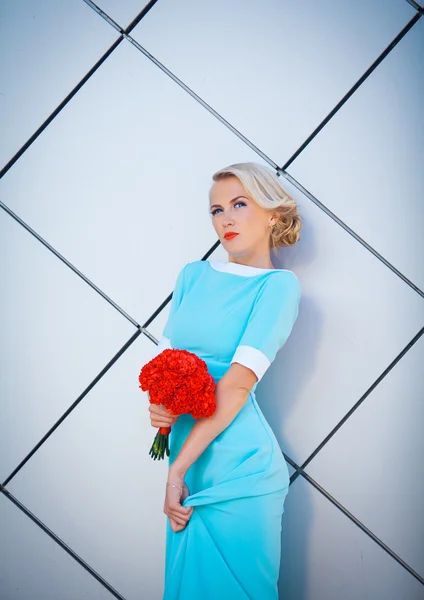 Daring blonde rebel with brightly painted lips. In a long blue dress with bouquet of red in hand. Against the background of the rectangular decorative walls.