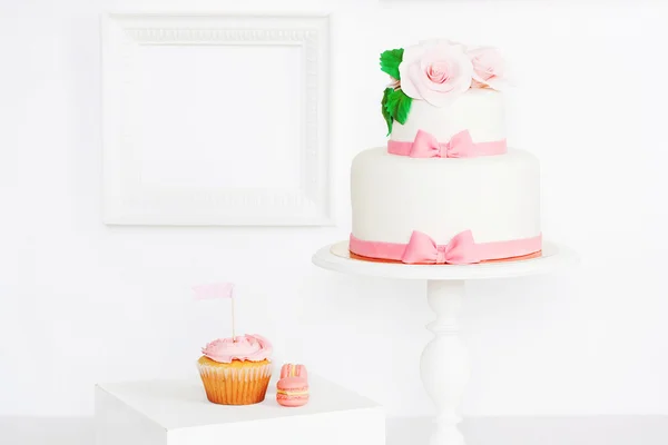 Cake decorated with roses and bows with cupcakes and macaroons standing on a table with frame
