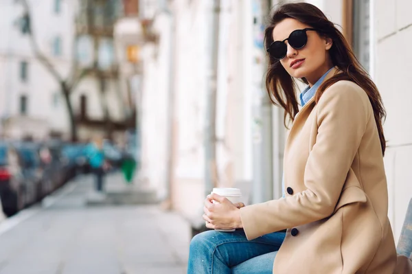 Beautiful and young girl in a coat and scarf and sunglasses sitting on the bench. Woman drinking coffee. Summer.
