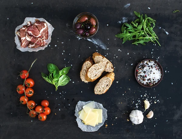 Ingredients for sandwich with smoked meat, baguette, basil, arugula, olives, cherry-tomatoes, parmesan cheese, garlic and spices over black grunge background