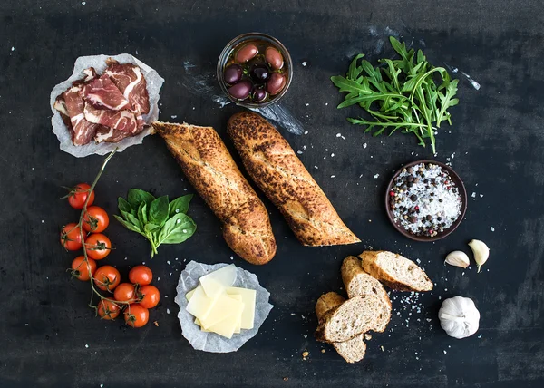Ingredients for sandwich with smoked meat, baguette, basil, arugula, olives, cherry-tomatoes, parmesan cheese, garlic and spices over black grunge background