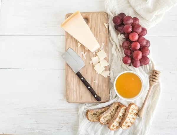 Parmesan cheese with grapes, honey and bread slices on wooden chopping board over rustic white background. Top view