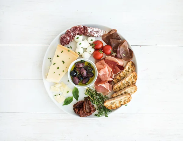 Wine appetizer set. Cherry-tomatoes, parmesan cheese, meat variety, bread slices, dried tomatoes, olives and basil on round ceramic plate over white wood backdrop