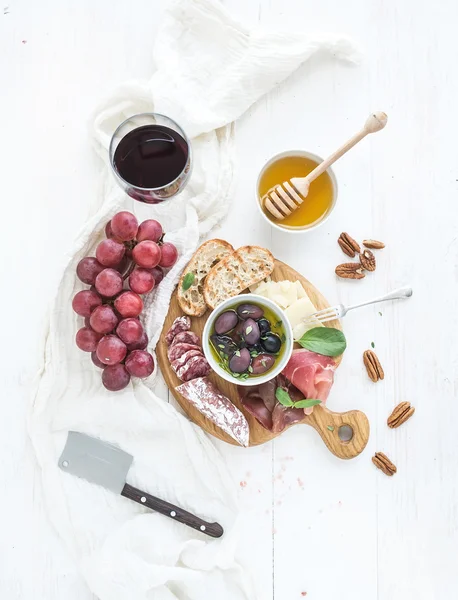 Wine appetizer set. Glass of red, grapes, parmesan cheese, meat variety, bread slices, pecan nuts, honey, olives and basil on rustic wooden board over white backdrop