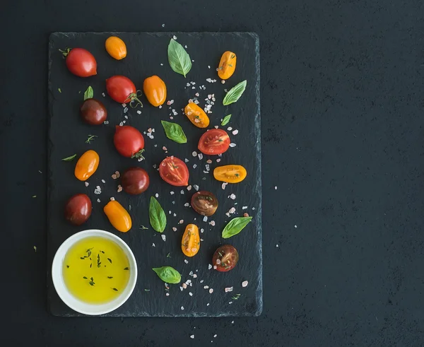 Cherry tomatoes of various color, basil leaves, spices and olive oil on black slate tray over dark grunge background. Top view