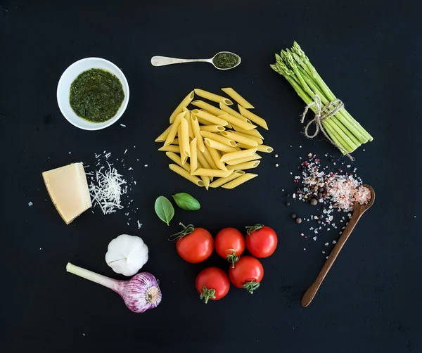 Ingredients for cooking pasta. Penne, green asparagus, basil, pesto sauce, garlic, spices, parmesan cheese and  cherry-tomatoes on dark grunge backdrop