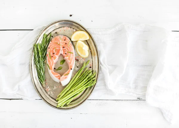 Raw salmon steak with asparagus, lemon, spices and rosemary on vintage silver tray over white wooden backdrop, top view