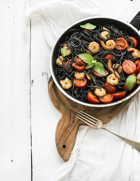 Black pasta spaghetti with shrimps, basil, pesto sauce and slow-roasted cherry-tomatoes in cooking pan on rustic chopping board over white wooden table
