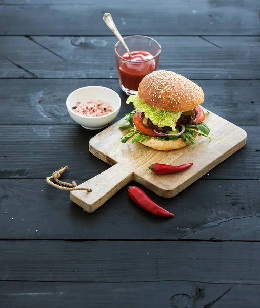 Fresh homemade burger on dark serving board with spicy tomato sauce, sea salt and herbs over black wooden background. Top view.