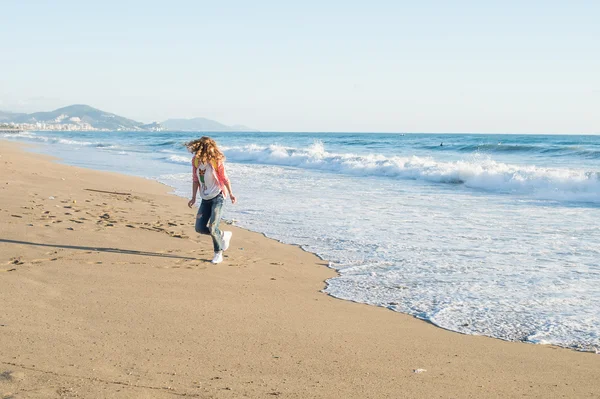 Young woman in red checkered shirt, jeans, white sneakers walking along beach and the stormy ocean on sunny winter day