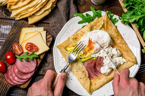 Rustic breakfast: crepe galette with meat, avocado, soft white cheese and poached egg on white plate