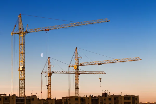 Construction cranes, building against blue sky with Moon at dawn
