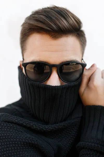 Close-up fashion portrait of a young handsome guy with stylish hairstyle in sunglasses and knitted sweater. Covers face with a cloth