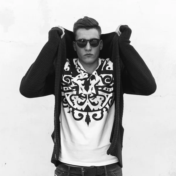 Black and white portrait of a young handsome guy in sunglasses and a sweater near a white wall