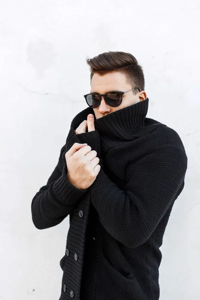 Young handsome guy with hair in a black sweater and sunglasses standing near a white wall.